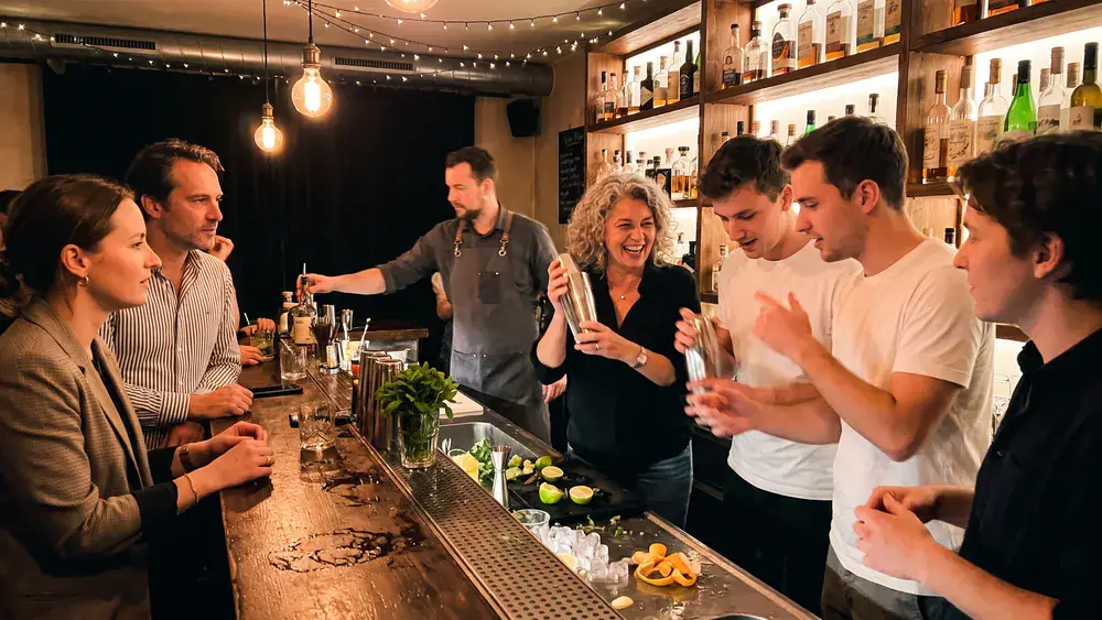 Cocktailkurs in Wien – Kollegen mixen Cocktails hinter einer beleuchteten Bar mit Anleitung in der Innenstadt